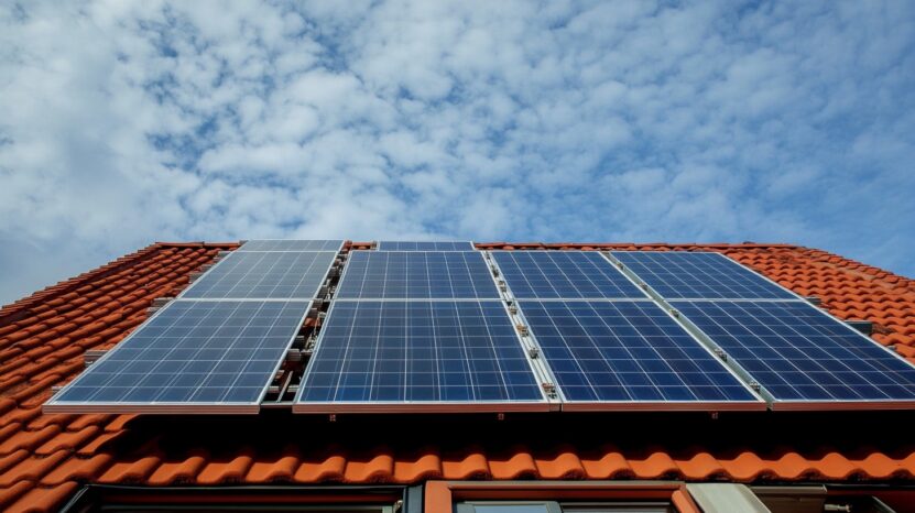 A close-up view of solar panels installed on a red-tiled roof under a partly cloudy sky. The panels are aligned neatly, capturing sunlight to generate clean, renewable energy. The bright sky and modern installation emphasize sustainability and efficient energy use.